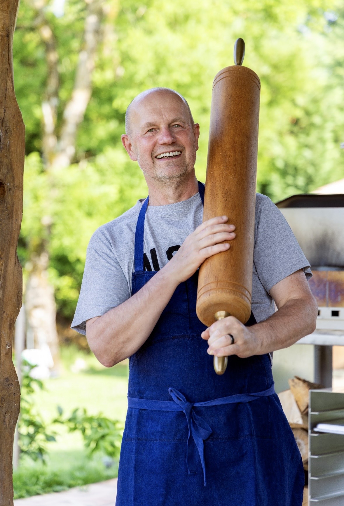 Chef Jacquy Pfeiffer smiling in blue apron holding wooden rolling pin